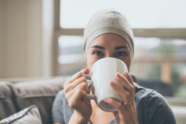 Junge Frau mit Krebs sitzt auf Couch und trinkt Tee.