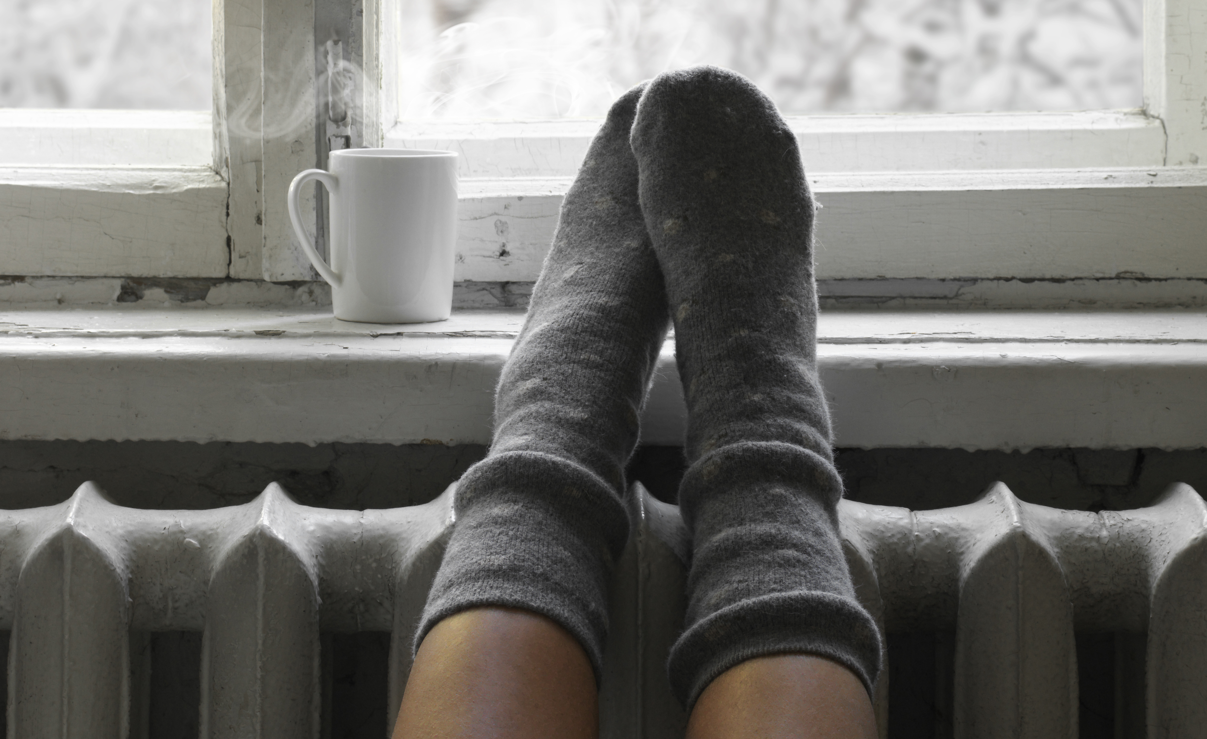 Füsse in warmen Socken liegen auf einer Heizung vor einem Fenster mit heissem Tee auf dem Fensterbrett.