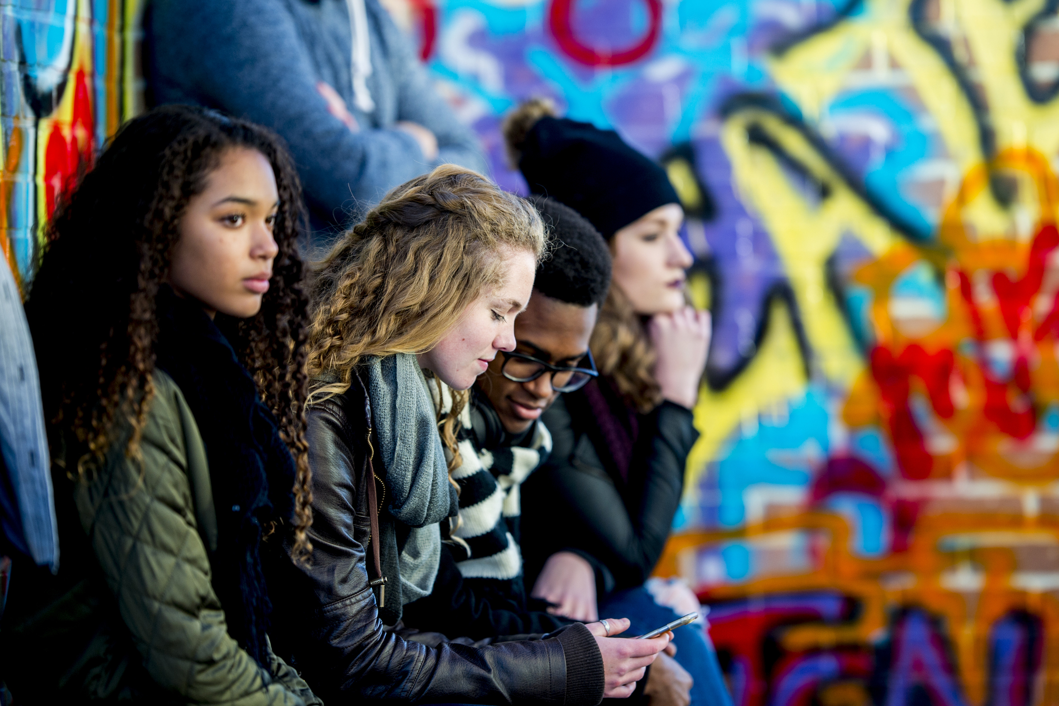 Eine Gruppe Jugendlicher sitzt vor einer Graffiti-Mauer und schaut ins Smartphone.