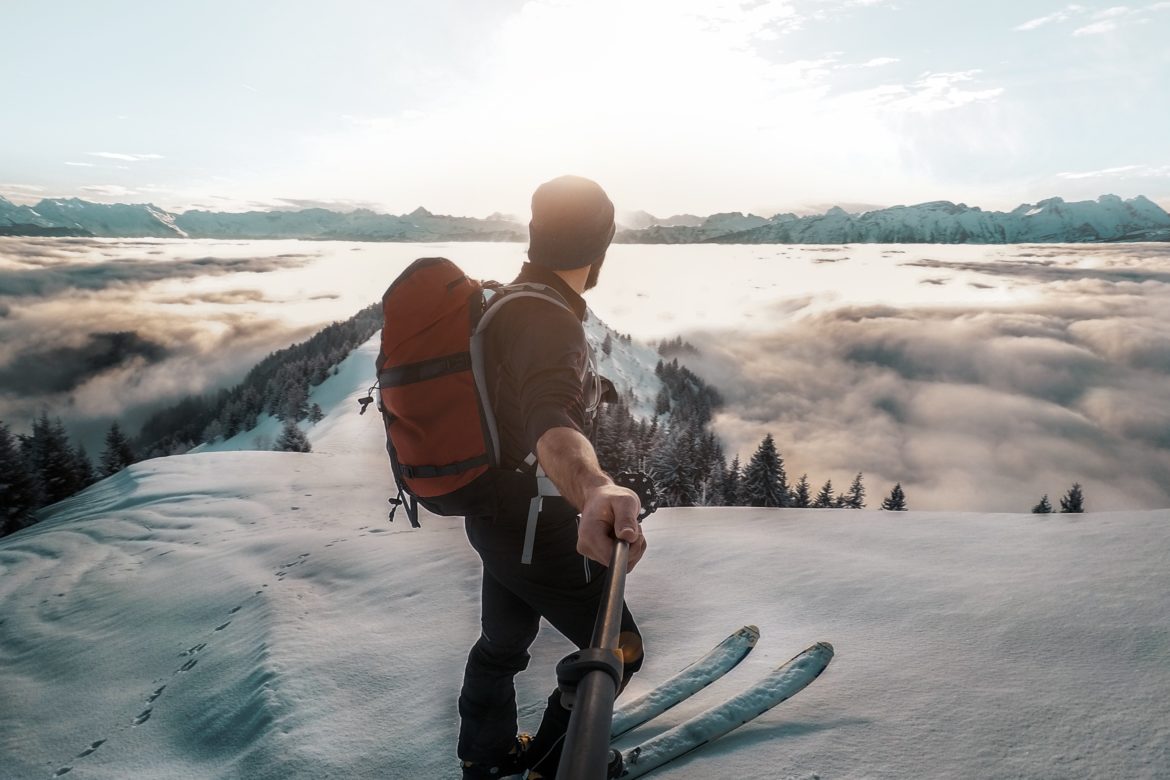 Mann auf Skiern macht Selfi mit Selfiestick vor verschneiter Bergkulisse.