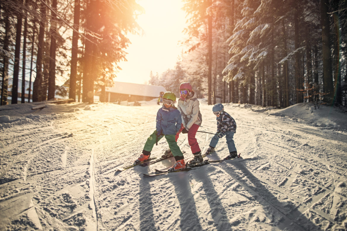 Drei Kinder fahren hintereinander auf einer verschneiten Piste und bei Sonne ski.