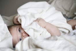 Ein Baby mit grossen blauen Augen liegt im Bett und hält eine weiche, weisse Decke vor sein Gesicht.