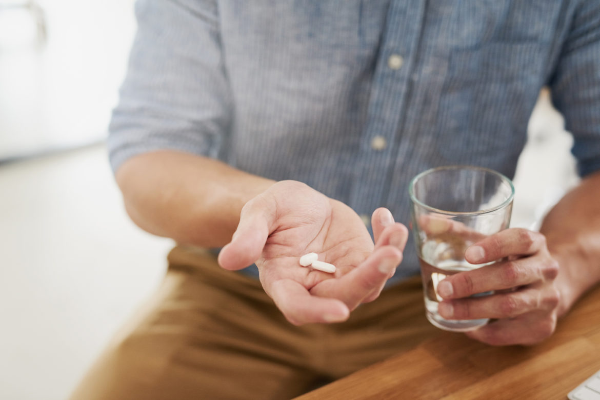 Mann in blauem Hemd und braunen Hosen mit Tabletten und einem Glas Wasser in der Hand.