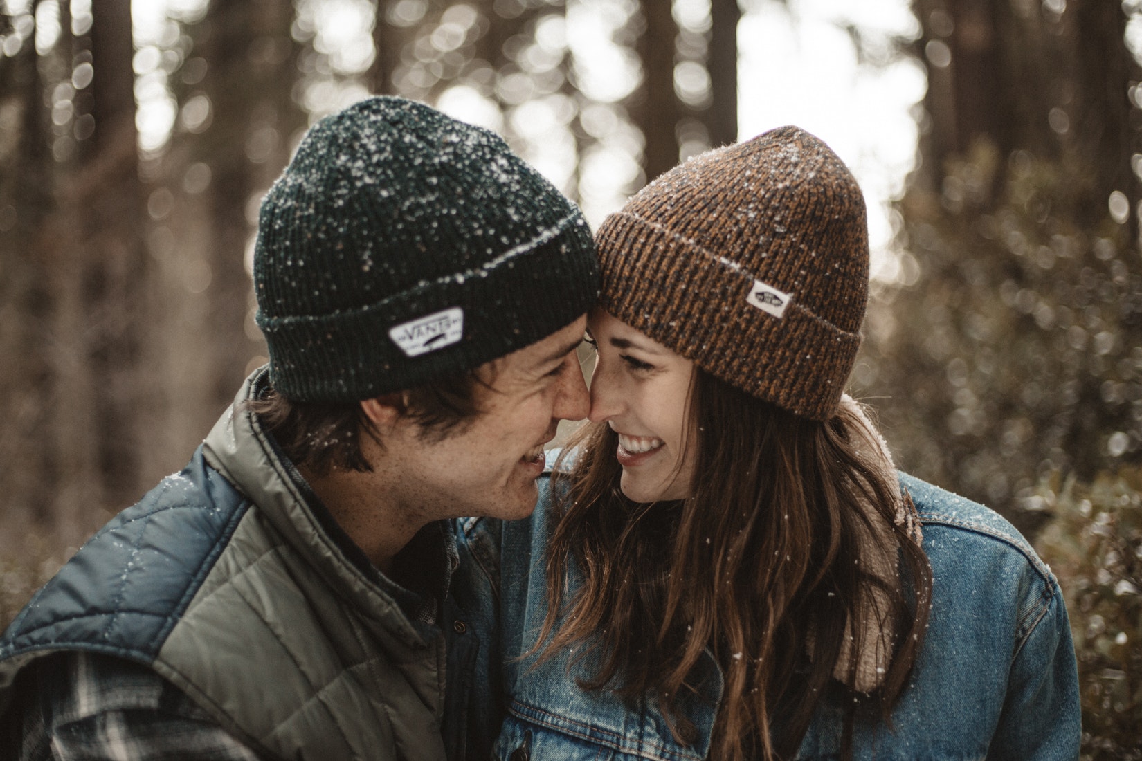 Junger Mann und junge Frau mit Mütze lachen sich Nase an Nase in Umarmung im Schnee an.