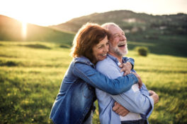Ein älteres Paar lachend sich umarmend in sonniger, grüner, hügeliger Landschaft.