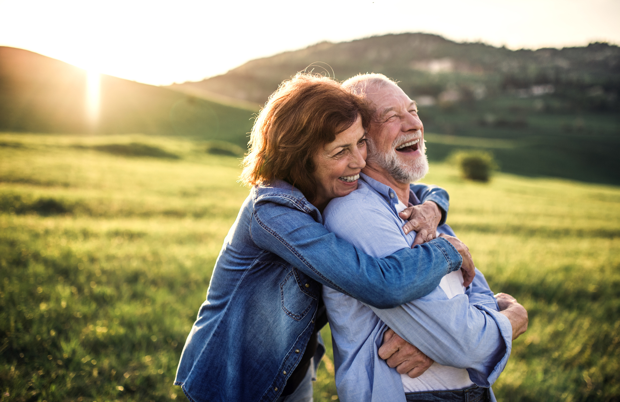 Ein älteres Paar lachend sich umarmend in sonniger, grüner, hügeliger Landschaft.