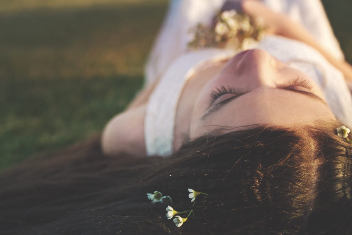 Junge Frau mit dunklen Haaren liegt mit geschlossenen AUgen schlafend auf einer Wiese mit Blumen in den Haaren und in der Hand.