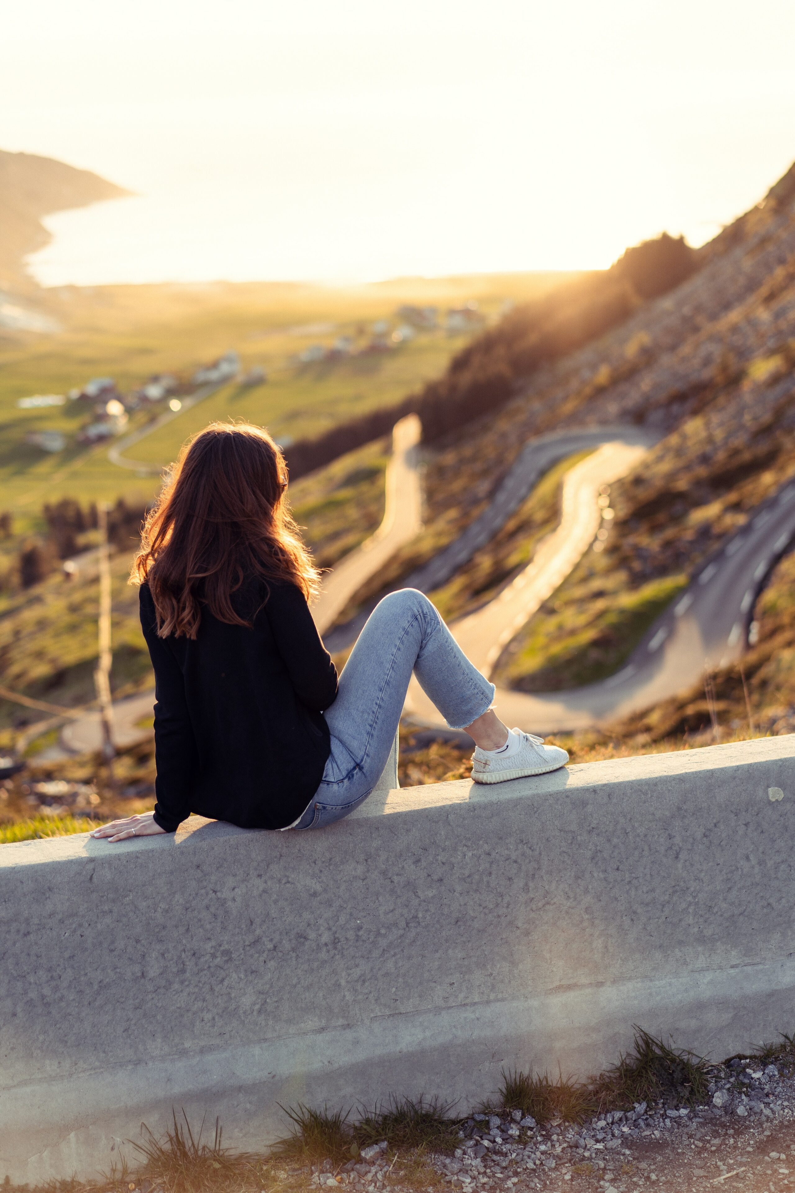 Junge Frau sitzt auf Mauer vor einer kurvigen Strasse