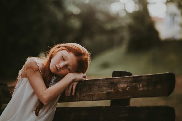Schlafstörungen Schulkind: Mädchen, das auf einer Bank sitzt und den Kopf auf den Armen abgelegt hat.