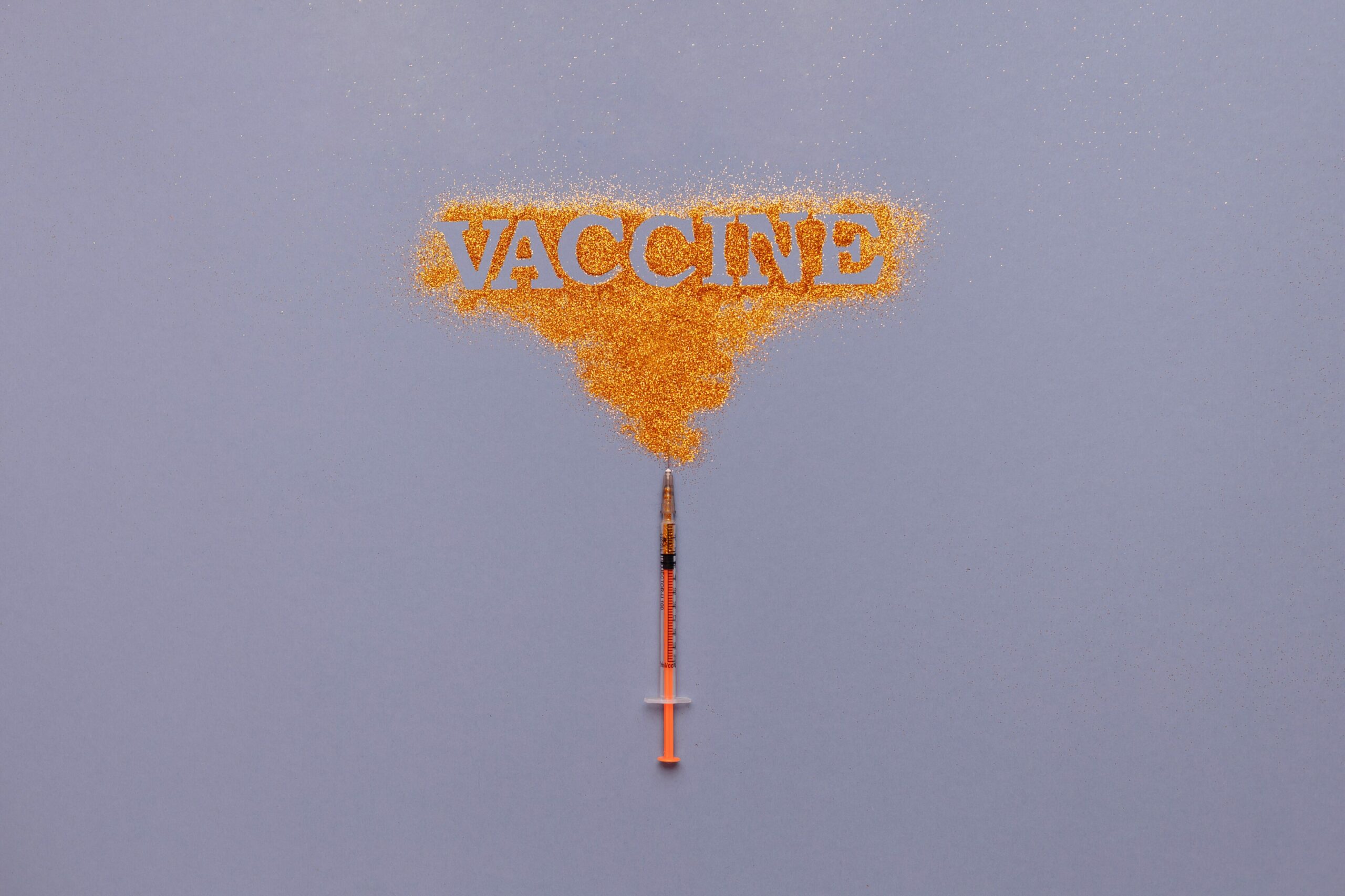 Grippeschutzimpfung: Auf violettem Grund liegt eine Spritze, die goldenen Glitzer verteilt, in dem "Vaccine" steht