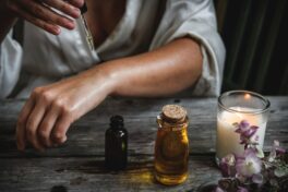 Ätherische Öle Anwendung: Eine Frau wendet Öle auf ihrem Unterarm an, auf dem Tisch stehen Ölflaschen und eine Kerze brennt.