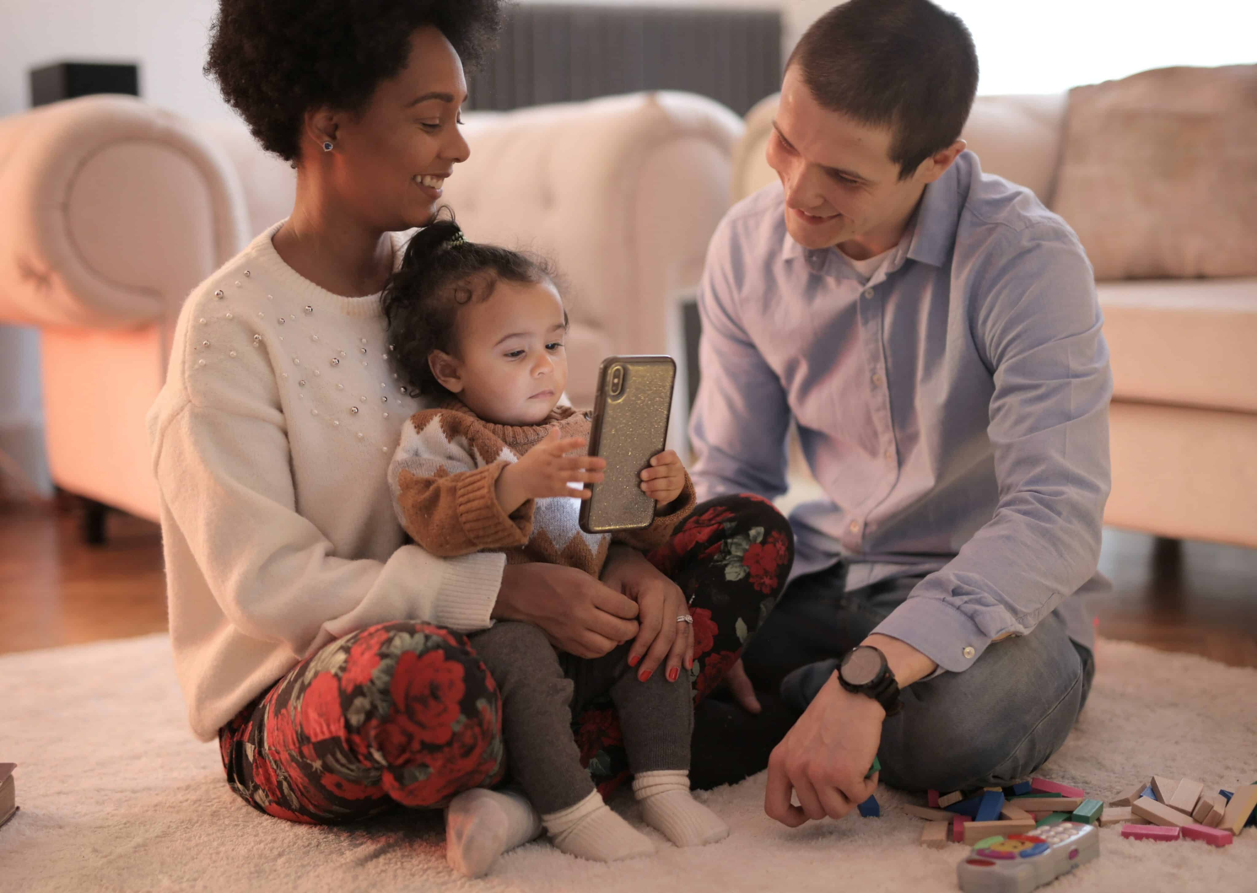 Kleinkinder Smartphone: Eltern sitzen mit ihrer kleinen Tochter auf dem Wohnzimmerboden und schauen auf den Bildschirm eines Smartphones.