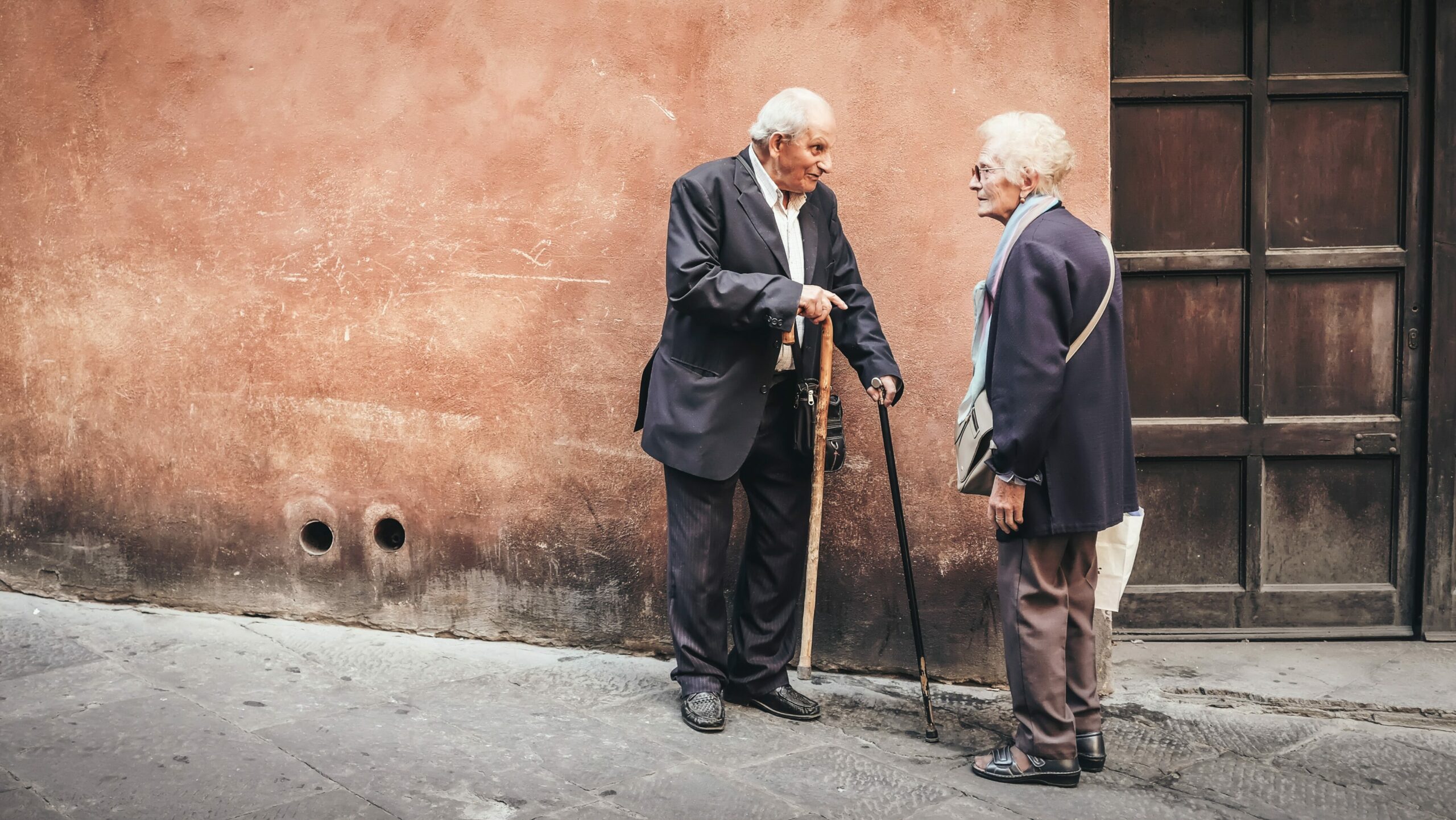 Stürze im Alter: Zwei ältere Menschen unterhalten sich vor einer roten Hauswand.