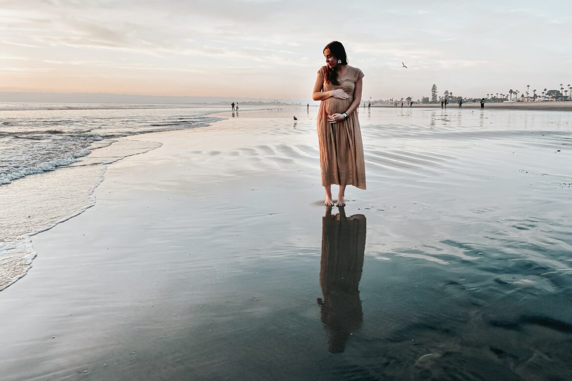 Kindheitstrauma Folgen: Schwangere Frau steht allein am Strand und hält ihren Bauch.