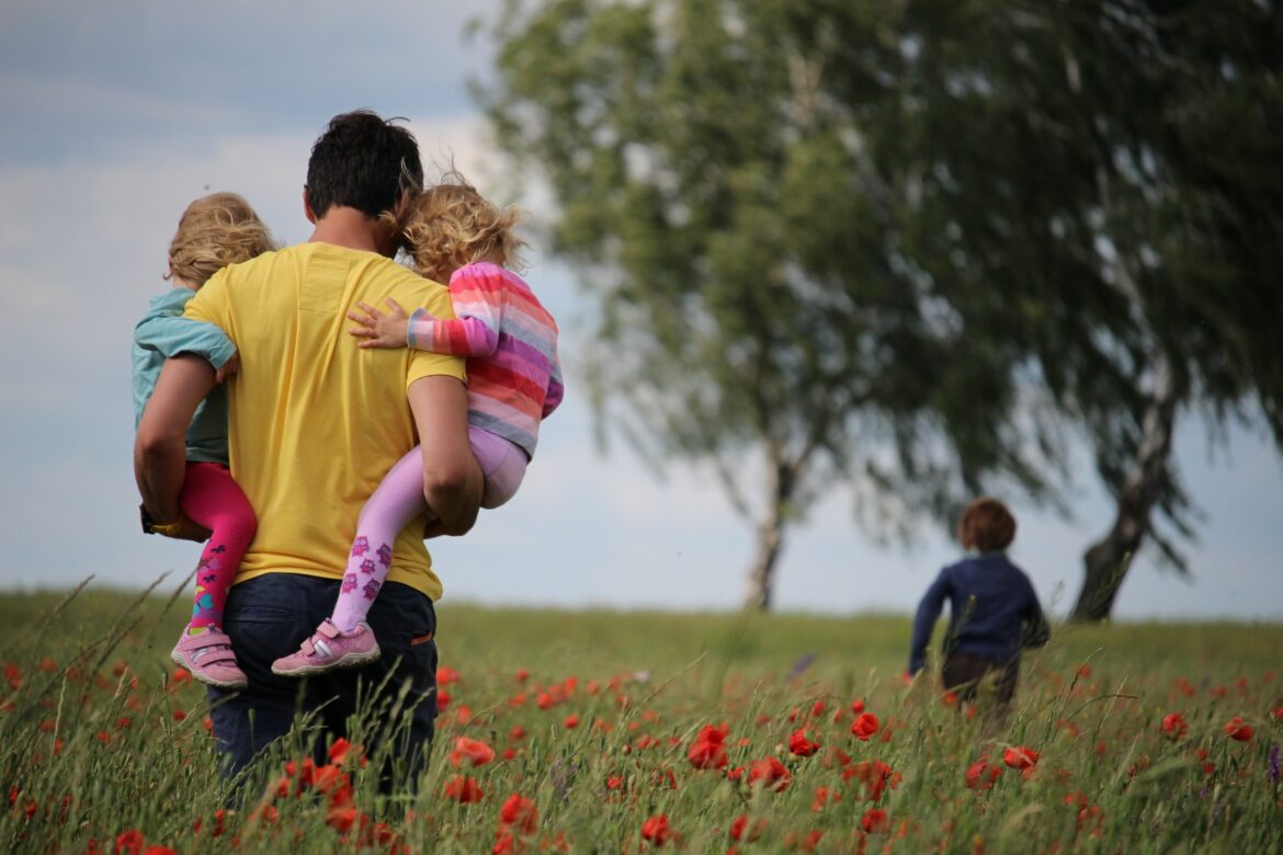 Krebsrisiko verringern: Vater trägt zwei Kinder über ein Feld, ein weiteres läuft voraus