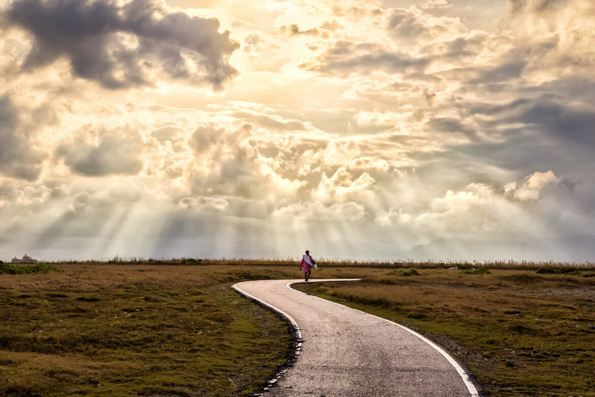 Diagnose Borderline: Ein Person geht alleine eine gewundene Strasse entlang, während die Sonnenstrahlen durch die Wolken auf sie scheinen.