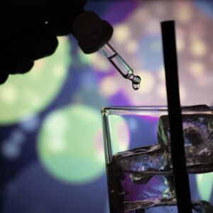 K.o.-Tropfen Schutz: Mit einer Pipette werden Drogen in ein Trinkglas mit Strohhalm und Eiswürfeln gegeben. Im Hintergrund bunte Lichter.