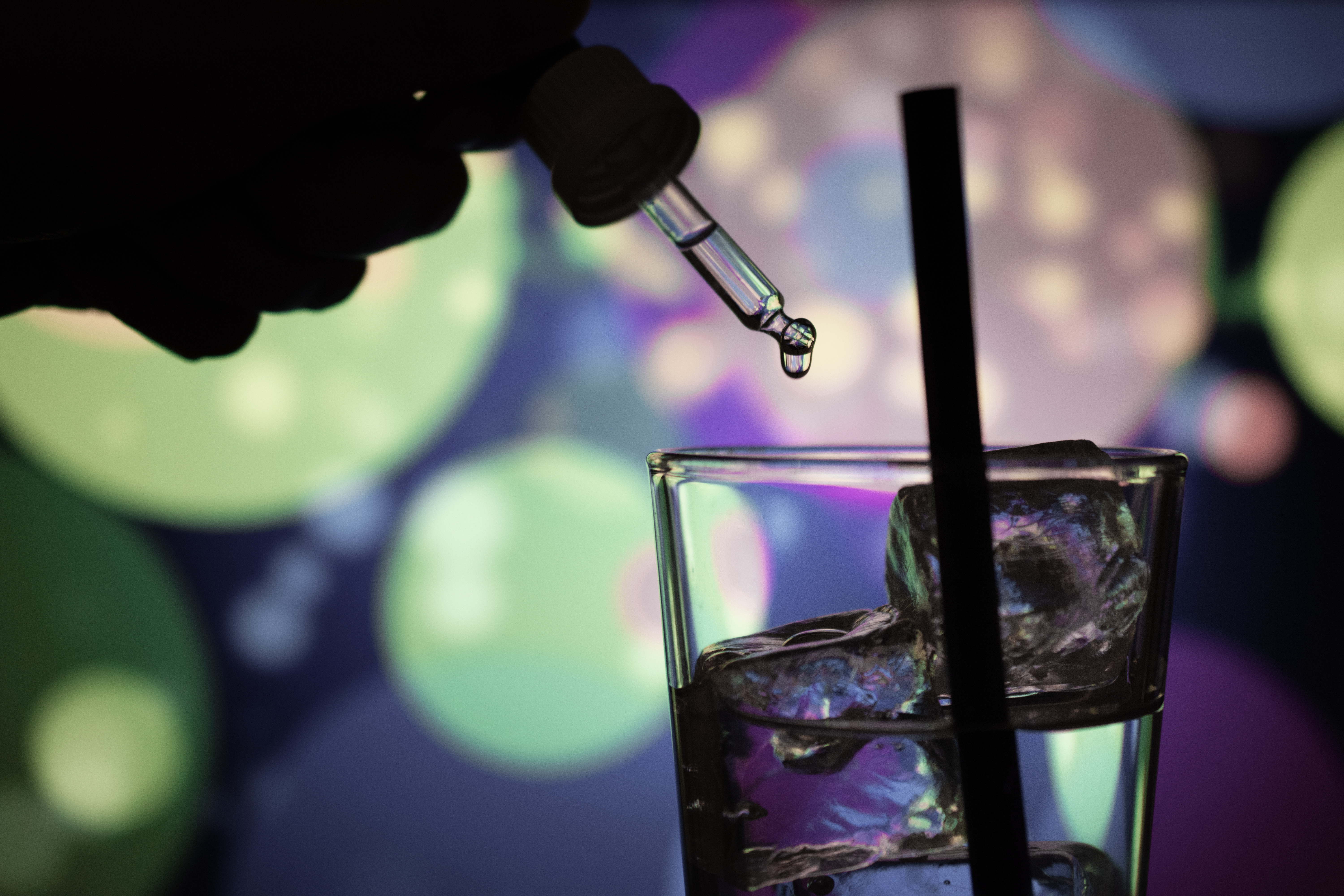K.o.-Tropfen Schutz: Mit einer Pipette werden Drogen in ein Trinkglas mit Strohhalm und Eiswürfeln gegeben. Im Hintergrund bunte Lichter.