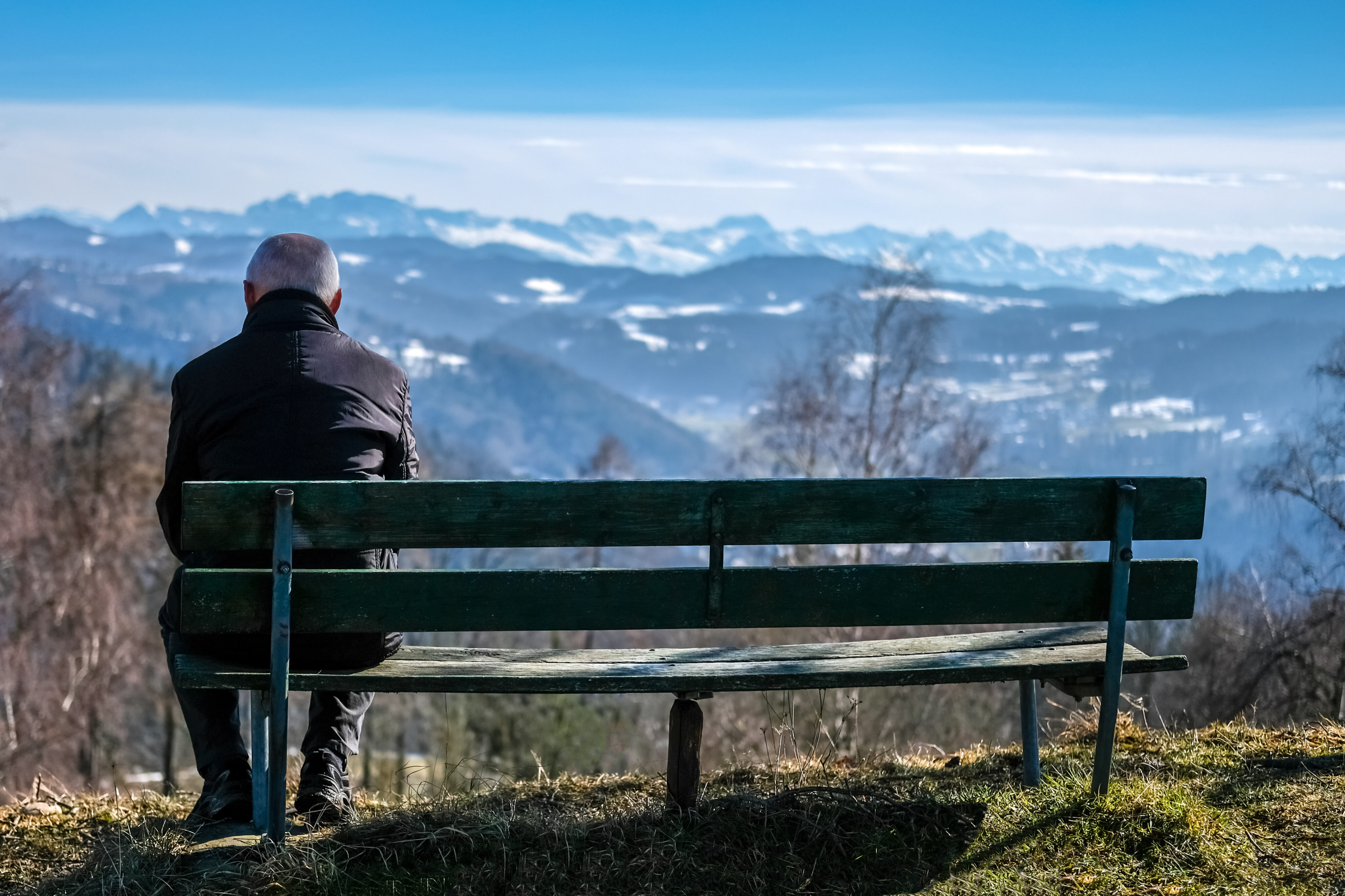 Einsam im Alter: Älterer Mann sitzt allein auf einer Bank und blickt auf eine Bergkulisse
