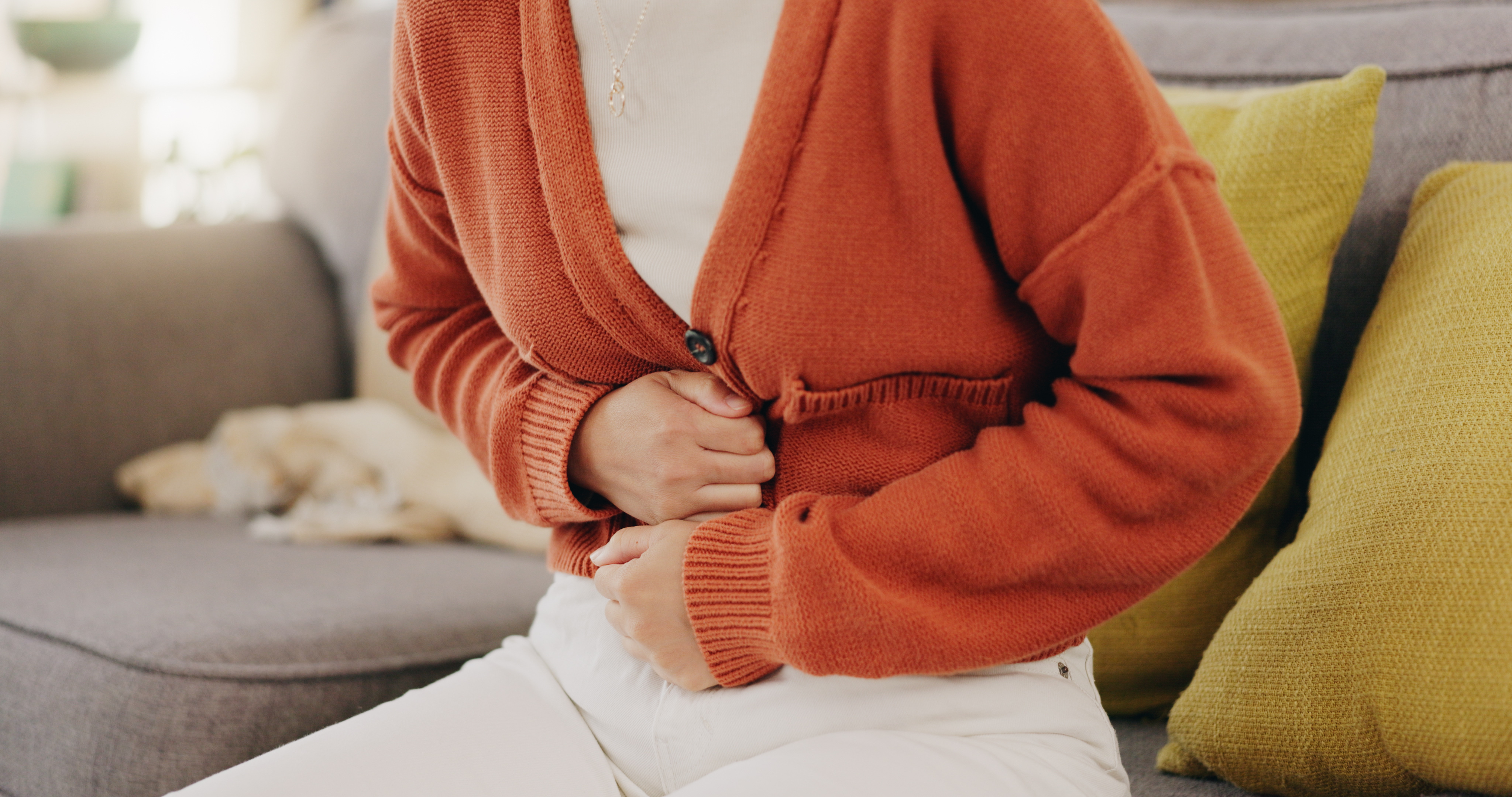 Ursachen für Blähbauch_Stomach ache, cramps and hands of woman with abdomen pain due to constipation, menstruation or ibs issue. Sick, home and person suffering and holding belly in a house lounge, couch and living room.