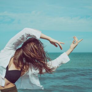Tanzen Übergewicht: Frau in Bewegung am Strand, gestikuliert tanzend mit ausgestreckten Armen gegen den Hintergrund des Meeres, ein inspirierendes Bild, das den positiven Einfluss von Tanzen auf die körperliche Aktivität und das Gewichtsmanagement hervorhebt.