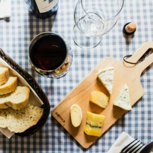 Ernährung Histamin:Käseplatte mit verschiedenen Sorten auf einem Holzbrett, frisches Brot im Korb und ein Glas Rotwein neben einer geöffneten Flasche auf einer blau-weiss karierten Tischdeck