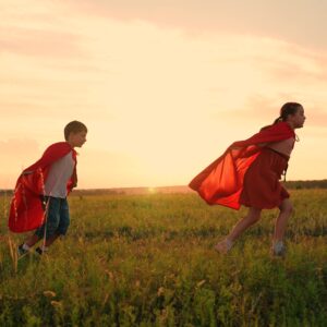 Verfrühte Pubertät: Drei Kinder rennen mit roten Umhängen im Sonnenuntergang über eine Wiese