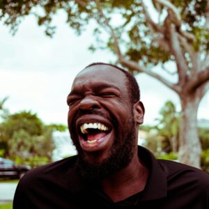 Entzündung Mundschleimhaut: Ein Mann lacht herzlich mit geschlossenen Augen vor einem Hintergrund mit grünen Bäumen