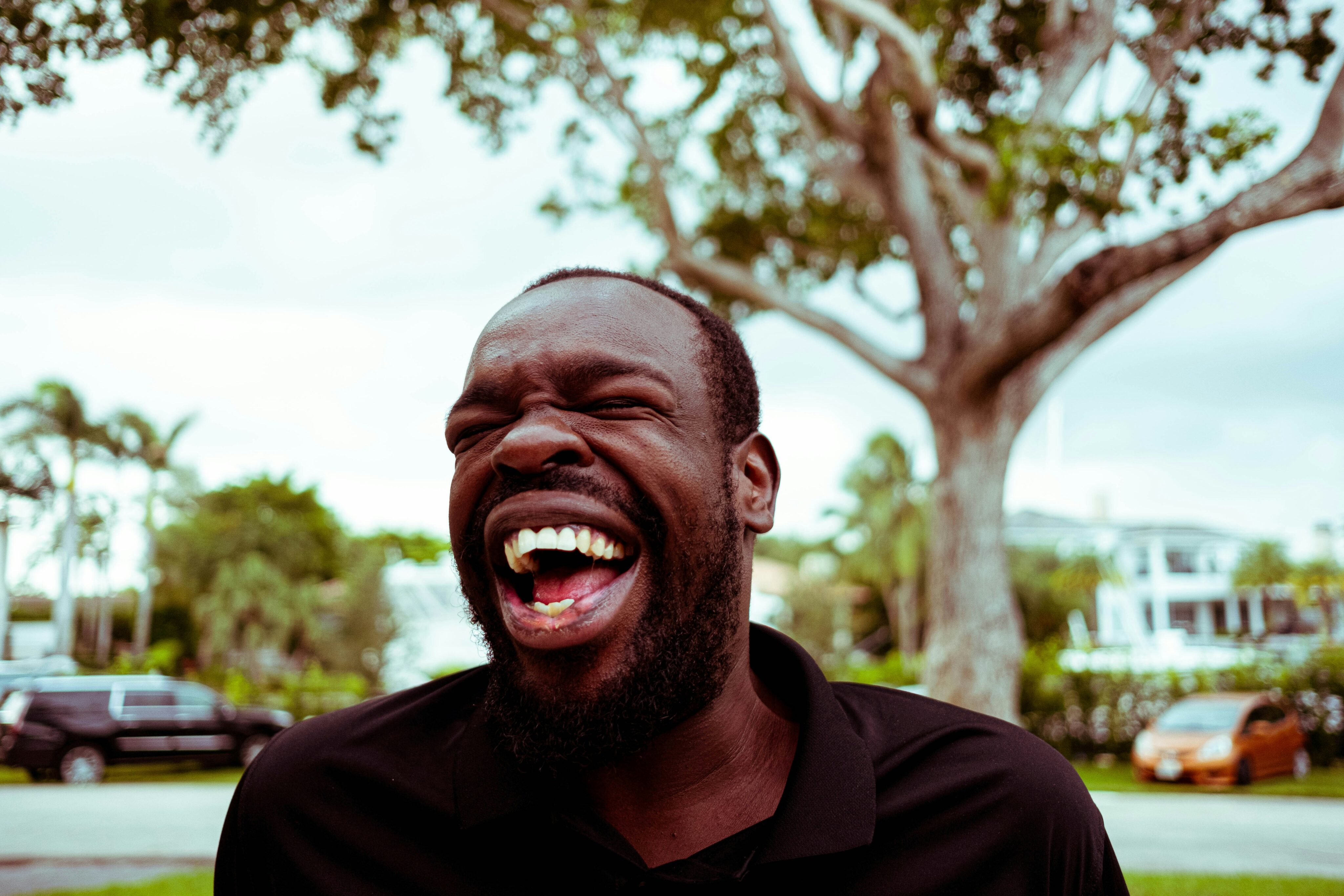 Entzündung Mundschleimhaut: Ein Mann lacht herzlich mit geschlossenen Augen vor einem Hintergrund mit grünen Bäumen